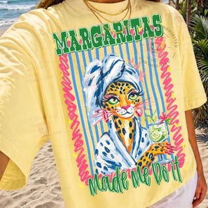 Pu&ograve; includere: T-shirt giallo con la scritta "Margaritas Made Me Do It". La grafica presenta un leopardo con un asciugamano e un cocktail. Il design include strisce blu e dettagli rosa.