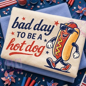 Puede incluir: Camiseta beige claro con la frase "bad day to be a hot dog" en azul marino y rojo. La camiseta presenta un perrito caliente de dibujos animados con gafas de sol caminando. La camiseta est&aacute; doblada sobre una manta azul.