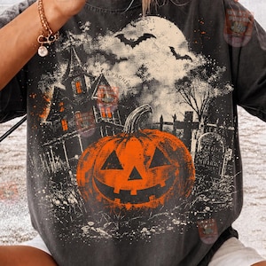 May include: A charcoal grey short-sleeve t-shirt featuring a graphic print of a large orange jack-o'-lantern in the foreground, a haunted house, a full moon, flying bats, and a tombstone in the background with a distressed, vintage-style texture.