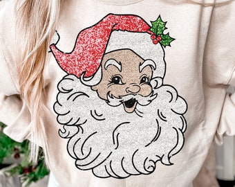 May include: A beige sweatshirt with a glittery Santa Claus design. The Santa Claus is wearing a red hat with a green holly sprig and red berries.