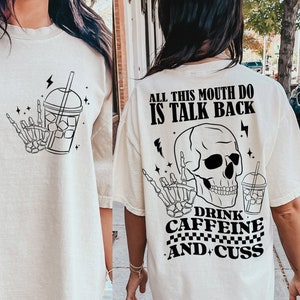 May include: A white oversized t-shirt with a black graphic design featuring a skull, a skeleton hand holding a cup, and the text "All this mouth do is talk back, Drink Caffeine and Cuss".