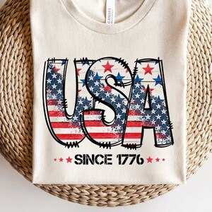 May include: A white t-shirt with a distressed graphic of the word "USA" in red, white, and blue with stars. The graphic is surrounded by stars and the text "Since 1776" is below the graphic.