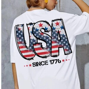 May include: White t-shirt with a graphic of the word "USA" in red, white, and blue with stars and stripes. The text "Since 1776" is printed below the graphic.