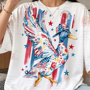 May include: White t-shirt with a quirky duck illustration. The duck is decorated with patriotic motifs, including the American flag and stars, and sports a matching hat. Red and blue brushstrokes and stars complete the design.