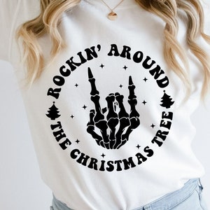 May include: A white long-sleeve sweatshirt featuring a black graphic of a skeleton hand making a rock-and-roll gesture. The design is encircled by the text "ROCKIN' AROUND THE CHRISTMAS TREE" with small star and pine tree icons.