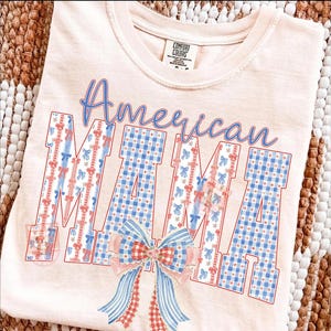 Op de afbeelding: Een lichtroze Comfort Colors T-shirt met de tekst "American MAMA" in blauwe letters en blokletters. De letters zijn versierd met rood, wit en blauw patronen, waaronder strikken en ruitjes. Onderaan zit een grote strik.