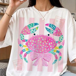 May include: White t-shirt featuring a colorful crab design. The crab is pink with blue, green, and yellow accents, and a pink bow. Vertical pink stripes are on the shirt. The shirt has the text "WHAT DESIGN".