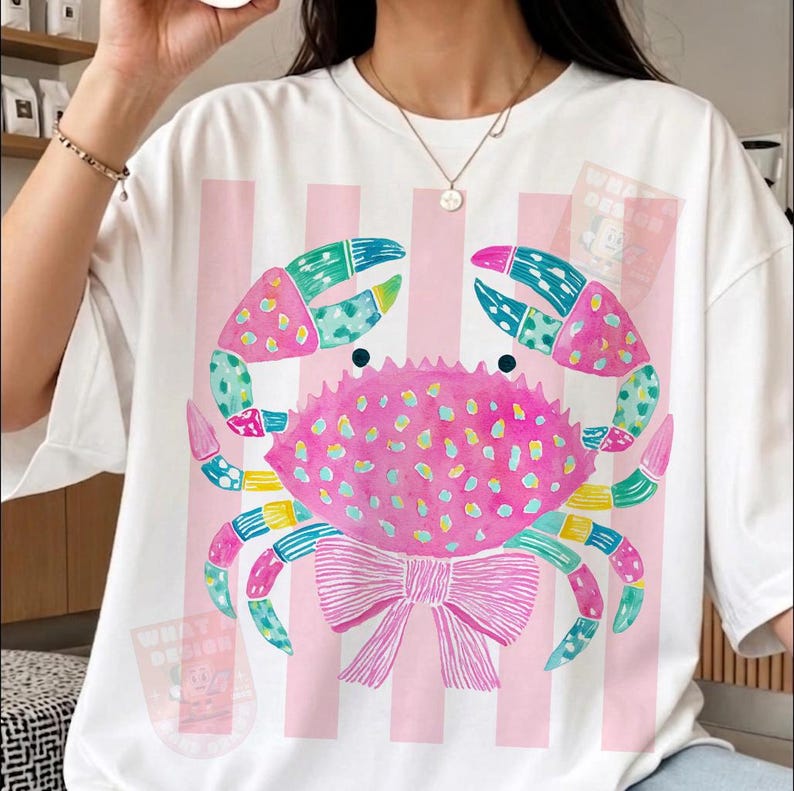 May include: White t-shirt featuring a colorful crab design. The crab is pink with blue, green, and yellow accents, and a pink bow. Vertical pink stripes are on the shirt. The shirt has the text "WHAT DESIGN".