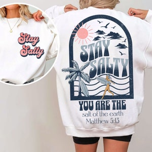 May include: White sweatshirt with a blue and white graphic on the back that says "Stay Salty" with a palm tree, sun, and a surfer. The text "You are the salt of the earth Matthew 5:13" is below the graphic. The front of the sweatshirt has the text "Stay Salty" in a retro font.