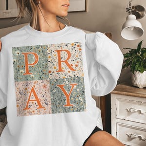 May include: A white long-sleeve sweatshirt featuring a large square graphic divided into four quadrants. Each quadrant displays a different floral pattern in muted tones, with the large orange letters P, R, A, and Y overlaid across the center.