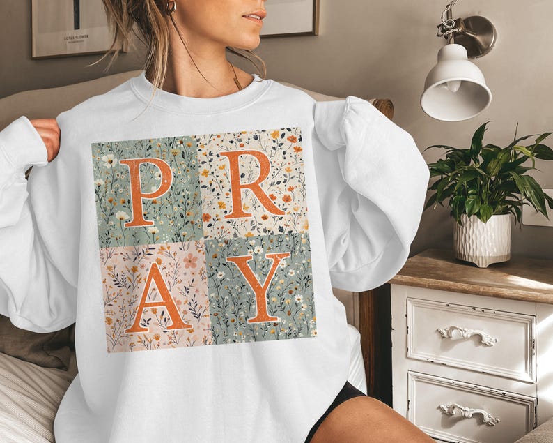May include: A white long-sleeve sweatshirt featuring a large square graphic divided into four quadrants. Each quadrant displays a different floral pattern in muted tones, with the large orange letters P, R, A, and Y overlaid across the center.