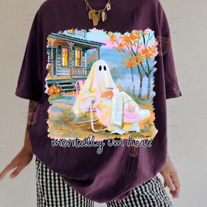 K&ouml;nnte beinhalten: Ein dunkelviolettes T-Shirt mit einer Grafik eines Geistes, der auf einem Stuhl vor einem Haus sitzt und einen Kaffeebecher h&auml;lt. Der Text "mentally i'm here" ist in Wei&szlig; unter der Illustration gedruckt, die orangefarbene B&auml;ume und K&uuml;rbisse zeigt.