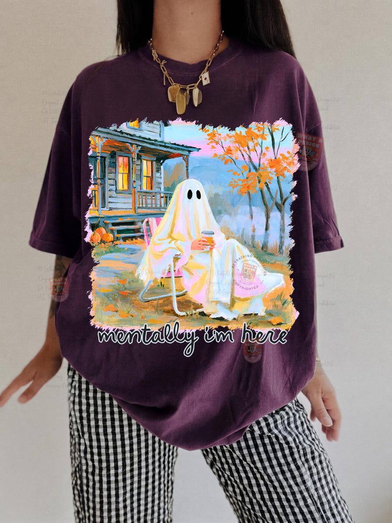 K&ouml;nnte beinhalten: Ein dunkelviolettes T-Shirt mit einer Grafik eines Geistes, der auf einem Stuhl vor einem Haus sitzt und einen Kaffeebecher h&auml;lt. Der Text "mentally i'm here" ist in Wei&szlig; unter der Illustration gedruckt, die orangefarbene B&auml;ume und K&uuml;rbisse zeigt.