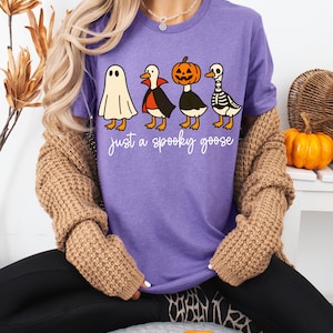 May include: A purple t-shirt featuring a Halloween-themed design with four cartoon geese dressed as a ghost, vampire, jack-o'-lantern, and skeleton. The text "just a spooky goose" is printed below the geese in a handwritten style.