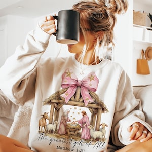 May include: A white sweatshirt with a pink bow and a nativity scene illustration. The text "The greatest gift of all" and "Matthew 1:21" are printed below the image.