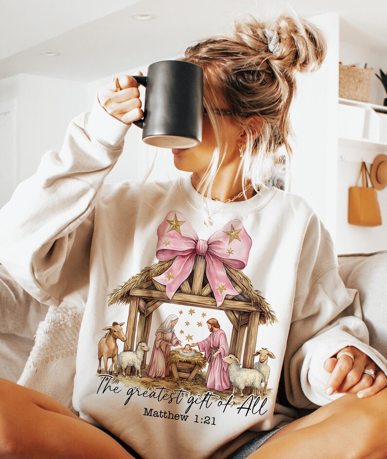May include: A white sweatshirt with a pink bow and a nativity scene illustration. The text "The greatest gift of all" and "Matthew 1:21" are printed below the image.