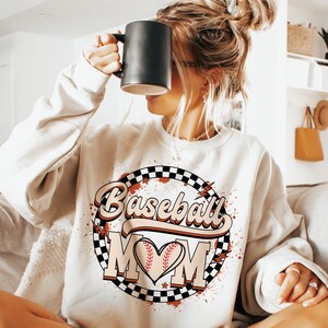 Peut inclure: Un sweat-shirt blanc avec un motif graphique repr&eacute;sentant un ballon de baseball dans un c&oelig;ur avec le texte "Baseball Mom" dans une police r&eacute;tro. Le graphique est entour&eacute; d'un cercle &agrave; carreaux avec des &eacute;claboussures rouges et blanches.