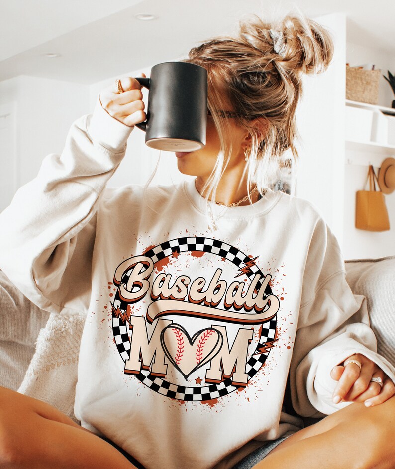 Peut inclure: Un sweat-shirt blanc avec un motif graphique repr&eacute;sentant un ballon de baseball dans un c&oelig;ur avec le texte "Baseball Mom" dans une police r&eacute;tro. Le graphique est entour&eacute; d'un cercle &agrave; carreaux avec des &eacute;claboussures rouges et blanches.