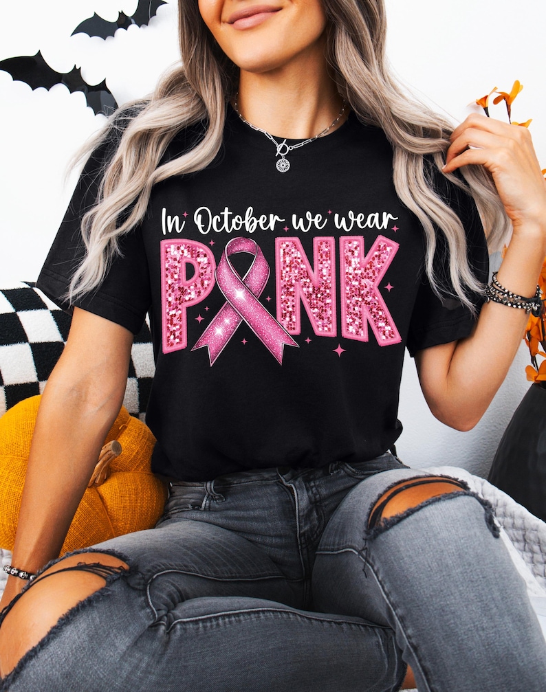 May include: A black t-shirt with a pink glitter design that says "In October we wear PINK" with a pink ribbon in the middle.