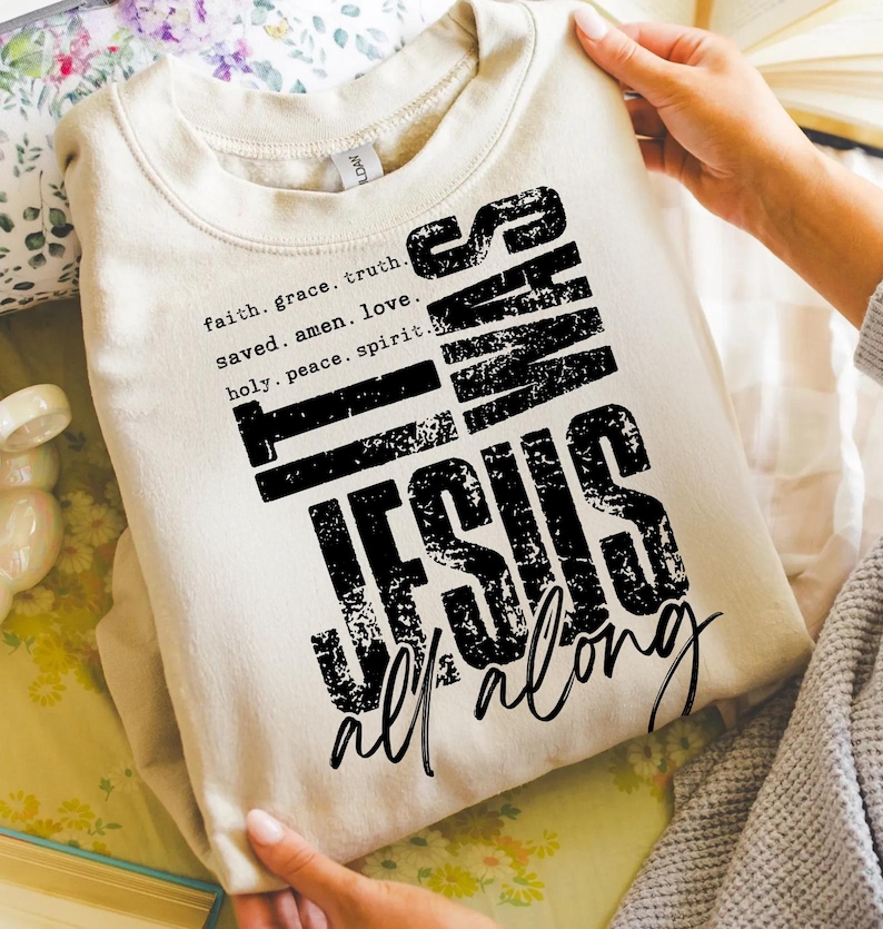 May include: A beige sweatshirt with the text "It Was Jesus All Along" in a distressed black font. The sweatshirt also features the words "faith, grace, truth, saved, amen, love, holy, peace, spirit" in a smaller font.