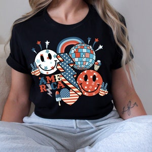 May include: Black t-shirt with a red, white and blue graphic design featuring a smiley face, a disco ball, fireworks, and the word "AMERICA".