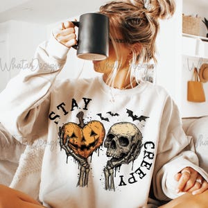Puede incluir: Sudadera crema con un gr&aacute;fico de Halloween. El dise&ntilde;o presenta un coraz&oacute;n de calabaza y una calavera sostenida por manos esquel&eacute;ticas, con las palabras "STAY CREEPY" y siluetas de murci&eacute;lagos. Se levanta una taza negra.