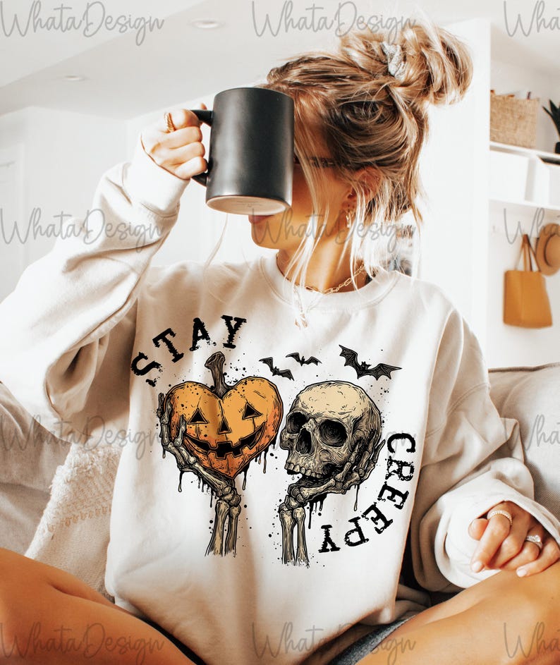 Puede incluir: Sudadera crema con un gr&aacute;fico de Halloween. El dise&ntilde;o presenta un coraz&oacute;n de calabaza y una calavera sostenida por manos esquel&eacute;ticas, con las palabras "STAY CREEPY" y siluetas de murci&eacute;lagos. Se levanta una taza negra.