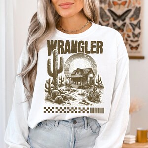 Pode incluir: Camisola branca de manga comprida com uma estampa gr&aacute;fica marrom de uma cena do deserto com uma casa, cactos e a palavra "WRANGLER" em letras negritas.