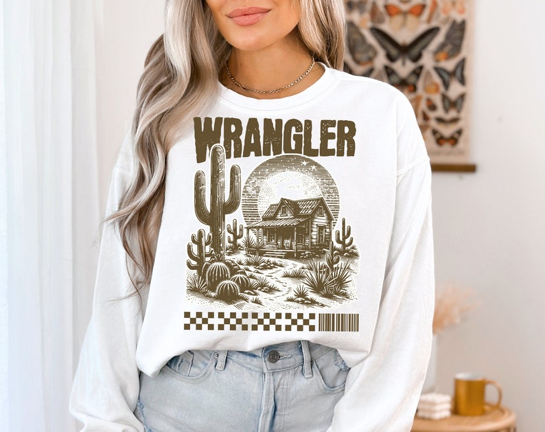 Pode incluir: Camisola branca de manga comprida com uma estampa gr&aacute;fica marrom de uma cena do deserto com uma casa, cactos e a palavra "WRANGLER" em letras negritas.