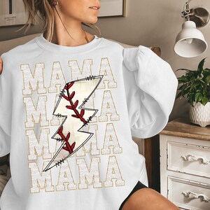 Peut inclure: Sweat-shirt blanc avec un graphique en paillettes dor&eacute;es d'un ballon de baseball cousu dans un &eacute;clair. Le texte "MAMA" est r&eacute;p&eacute;t&eacute; quatre fois en paillettes dor&eacute;es.