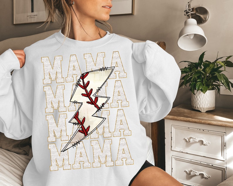 Peut inclure: Sweat-shirt blanc avec un graphique en paillettes dor&eacute;es d'un ballon de baseball cousu dans un &eacute;clair. Le texte "MAMA" est r&eacute;p&eacute;t&eacute; quatre fois en paillettes dor&eacute;es.