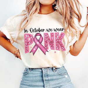 May include: A white t-shirt with the text "In October we wear PINK" in pink glitter letters with a pink ribbon in the center.