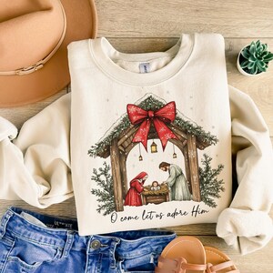 May include: Cream-colored sweatshirt featuring a Christmas nativity scene with a red bow and the text "O come let us adore Him." The design includes a wooden stable, figures, and evergreen accents. The sweatshirt is paired with blue jeans and a tan hat.