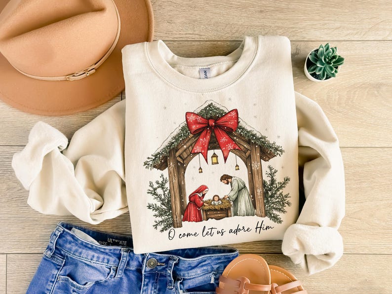 May include: Cream-colored sweatshirt featuring a Christmas nativity scene with a red bow and the text "O come let us adore Him." The design includes a wooden stable, figures, and evergreen accents. The sweatshirt is paired with blue jeans and a tan hat.