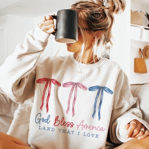 May include: A white sweatshirt with a red, white and blue bow design and the text "God Bless America Land That I Love".
