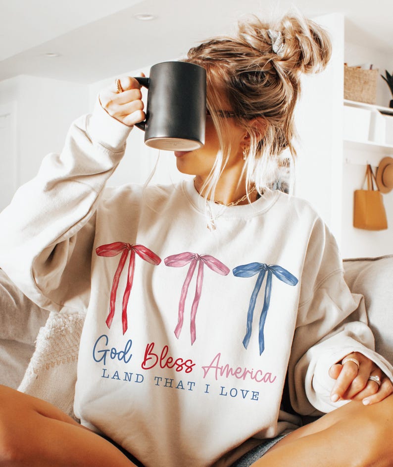 May include: A white sweatshirt with a red, white and blue bow design and the text "God Bless America Land That I Love".