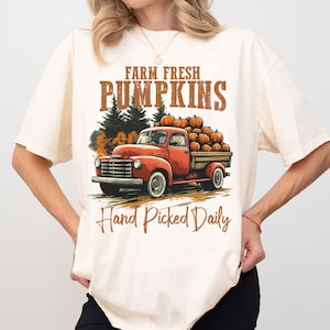 Op de afbeelding: Een wit T-shirt met een vintage-stijl afbeelding van een rode vrachtwagen die pompoenen vervoert. De tekst op het shirt luidt "Farm Fresh Pumpkins Hand Picked Daily".