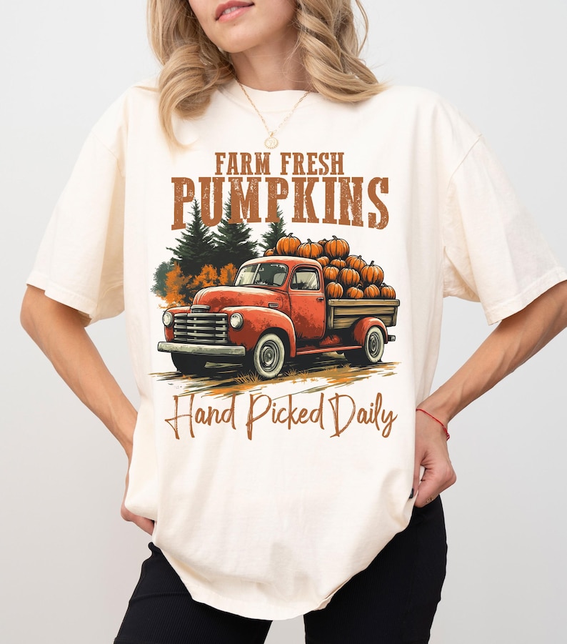 Op de afbeelding: Een wit T-shirt met een vintage-stijl afbeelding van een rode vrachtwagen die pompoenen vervoert. De tekst op het shirt luidt "Farm Fresh Pumpkins Hand Picked Daily".