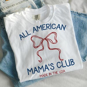 May include: White t-shirt with the text "ALL AMERICAN MAMA'S CLUB" in navy blue and a red bow graphic. The words "MADE IN THE USA" are printed at the bottom. The shirt is folded on top of a pair of blue jeans.
