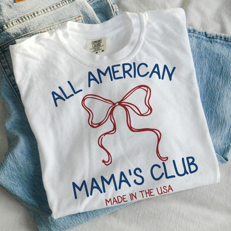 May include: White t-shirt with the text "ALL AMERICAN MAMA'S CLUB" in navy blue and a red bow graphic. The words "MADE IN THE USA" are printed at the bottom. The shirt is folded on top of a pair of blue jeans.