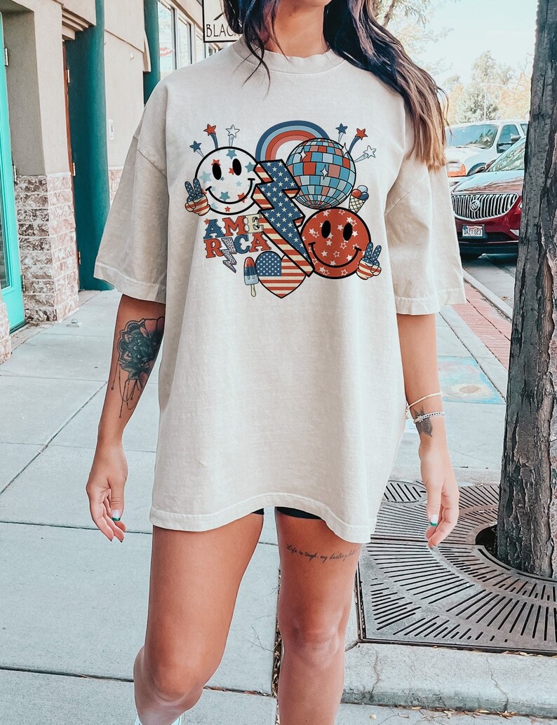 May include: An oversized beige t-shirt featuring a graphic print of a red, white and blue American flag, smiley faces and a disco ball. The text "AMERICA" is printed on the shirt.