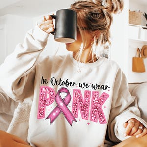 May include: White sweatshirt with the text "In October we wear PINK" in black lettering. The word "PINK" is in pink glitter letters with a pink ribbon in the center.