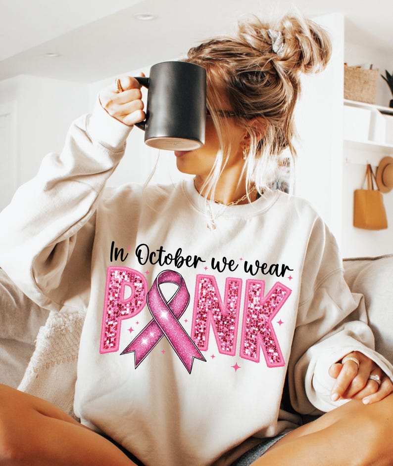 May include: White sweatshirt with the text "In October we wear PINK" in black lettering. The word "PINK" is in pink glitter letters with a pink ribbon in the center.
