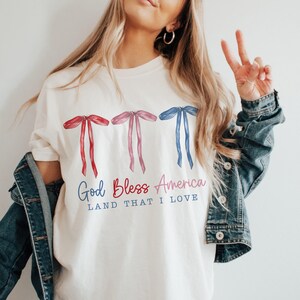 May include: White t-shirt with a red, white and blue bow design and the text "God Bless America Land That I Love".