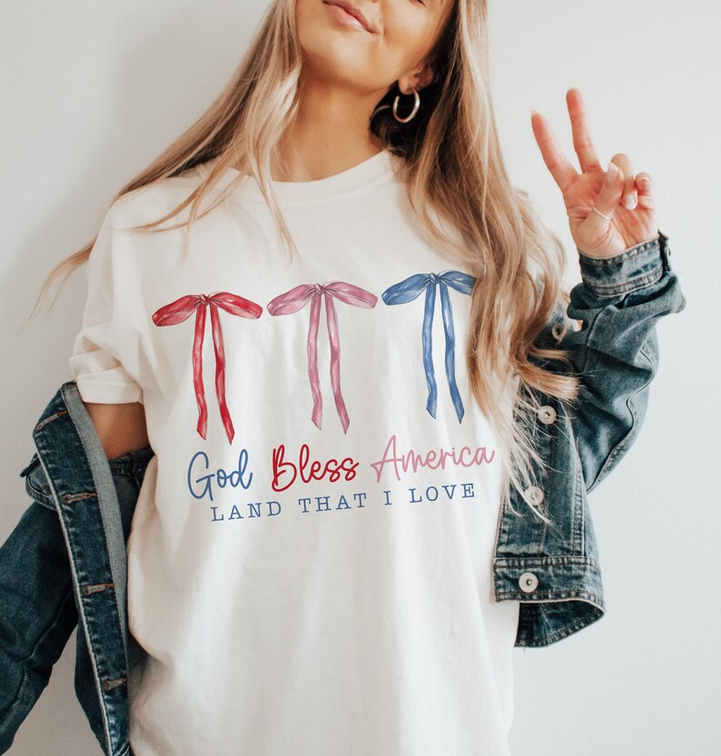 May include: White t-shirt with a red, white and blue bow design and the text "God Bless America Land That I Love".