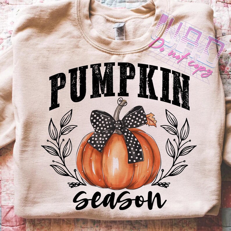 May include: A beige sweatshirt with a graphic featuring a pumpkin with a black polka-dot bow, surrounded by leaves. The words "PUMPKIN SEASON" are printed above and below the pumpkin in a bold, black font. A fall-themed design.