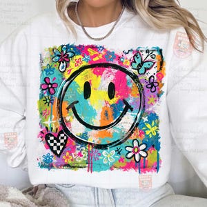 May include: White sweatshirt featuring a colorful, abstract smiley face design. The design includes a black smiley face, flowers, a butterfly, and a checkered heart, all set against a vibrant, multi-colored background.
