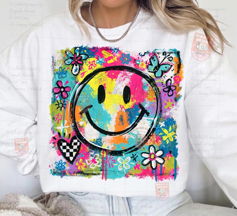 May include: White sweatshirt featuring a colorful, abstract smiley face design. The design includes a black smiley face, flowers, a butterfly, and a checkered heart, all set against a vibrant, multi-colored background.
