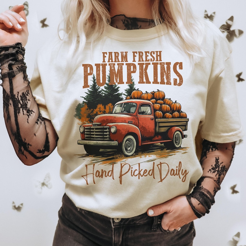 Op de afbeelding: Een wit T-shirt met een vintage-stijl afbeelding van een rode pick-up truck vol met pompoenen. De tekst "Farm Fresh Pumpkins Hand Picked Daily" is op het shirt gedrukt.