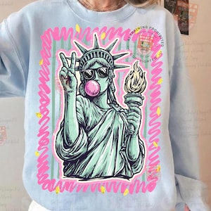 Puede incluir: Sudadera azul claro con un gr&aacute;fico de la Estatua de la Libertad con gafas de sol y soplando una burbuja. La estatua sostiene una antorcha y hace el signo de la paz. El dise&ntilde;o est&aacute; enmarcado en rosa con detalles amarillos.